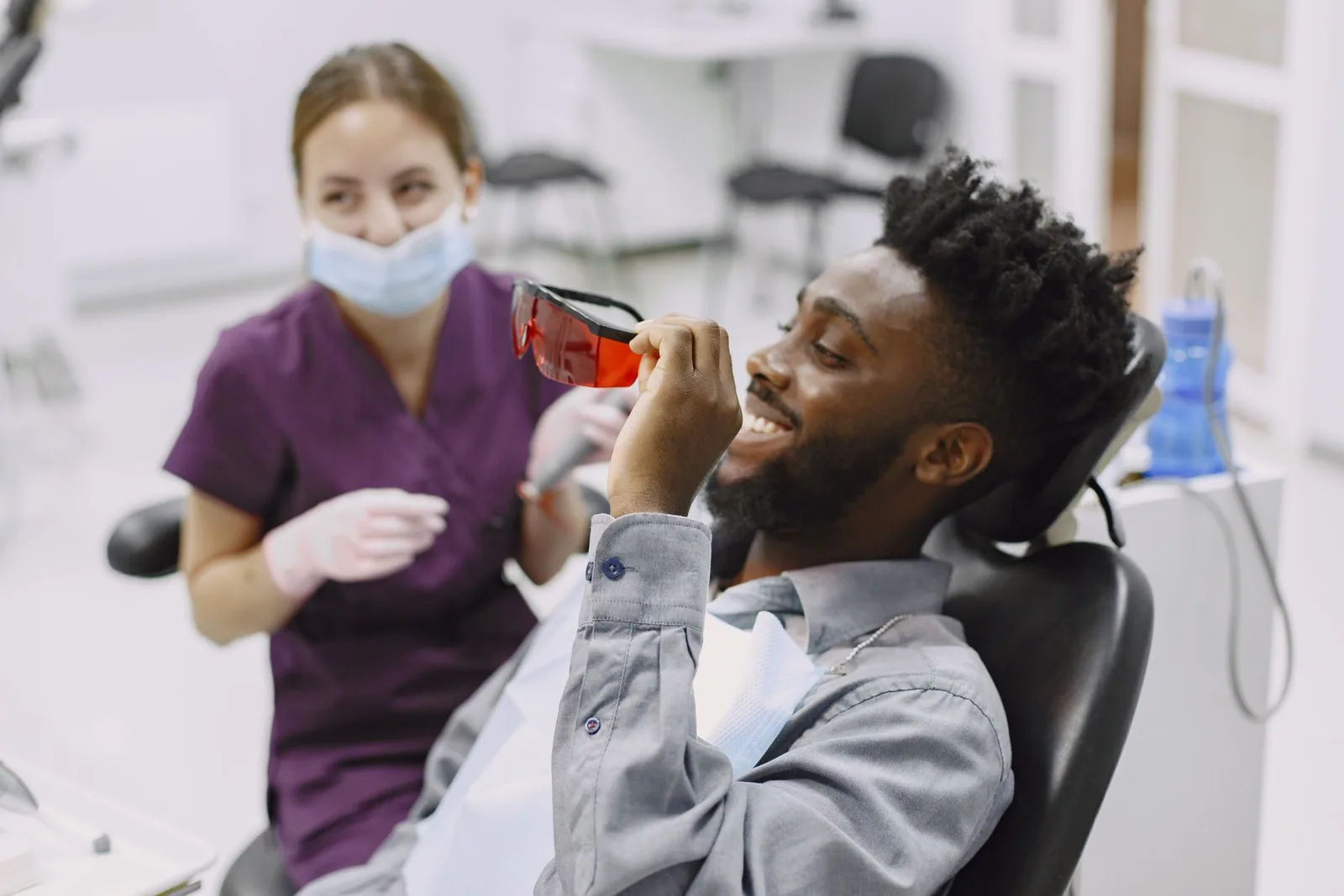 A smiling patient holds protective glasses while seated in a dental treatment room